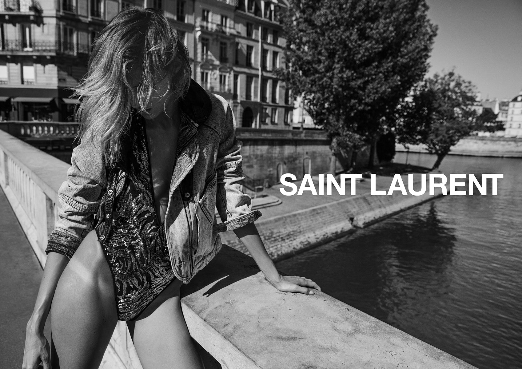 Person leaning on a bridge with water and buildings in the background, featuring the Saint Laurent logo.